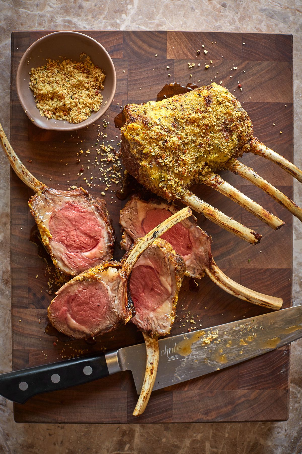 On a wooden cutting board, a rack of lamb crusted with bread crumbs is being cut by a large knife.