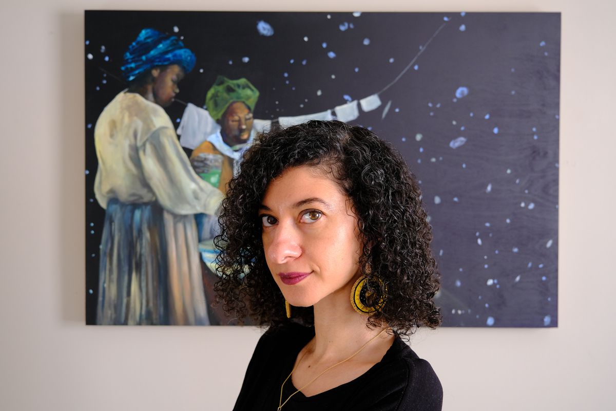 The physicist Chanda Prescod-Weinstein looks at the camera while standing in front of a painting depicting two dark-skinned women hanging laundry in front of a field of stars.