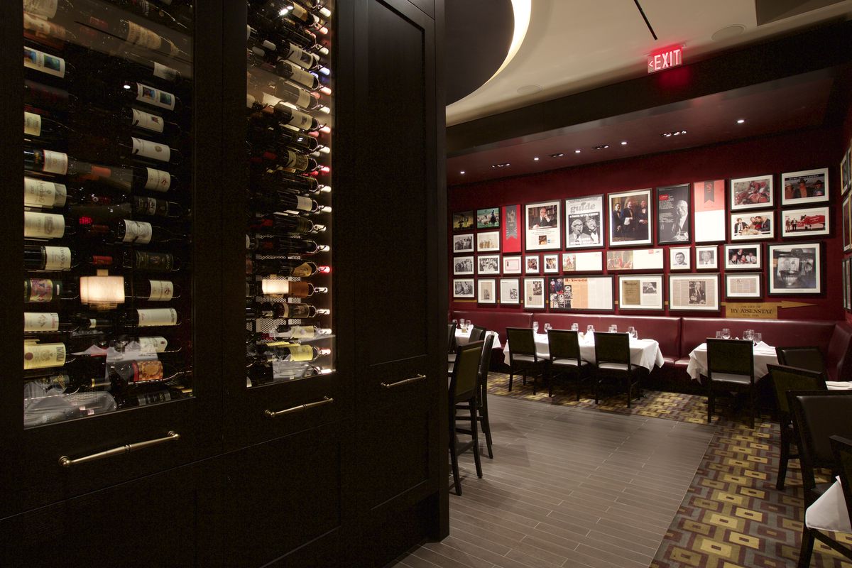 A large wine vault with glass windows displaying bottles, which curves toward a seating area set beneath a wall of memorabilia and news clippings.