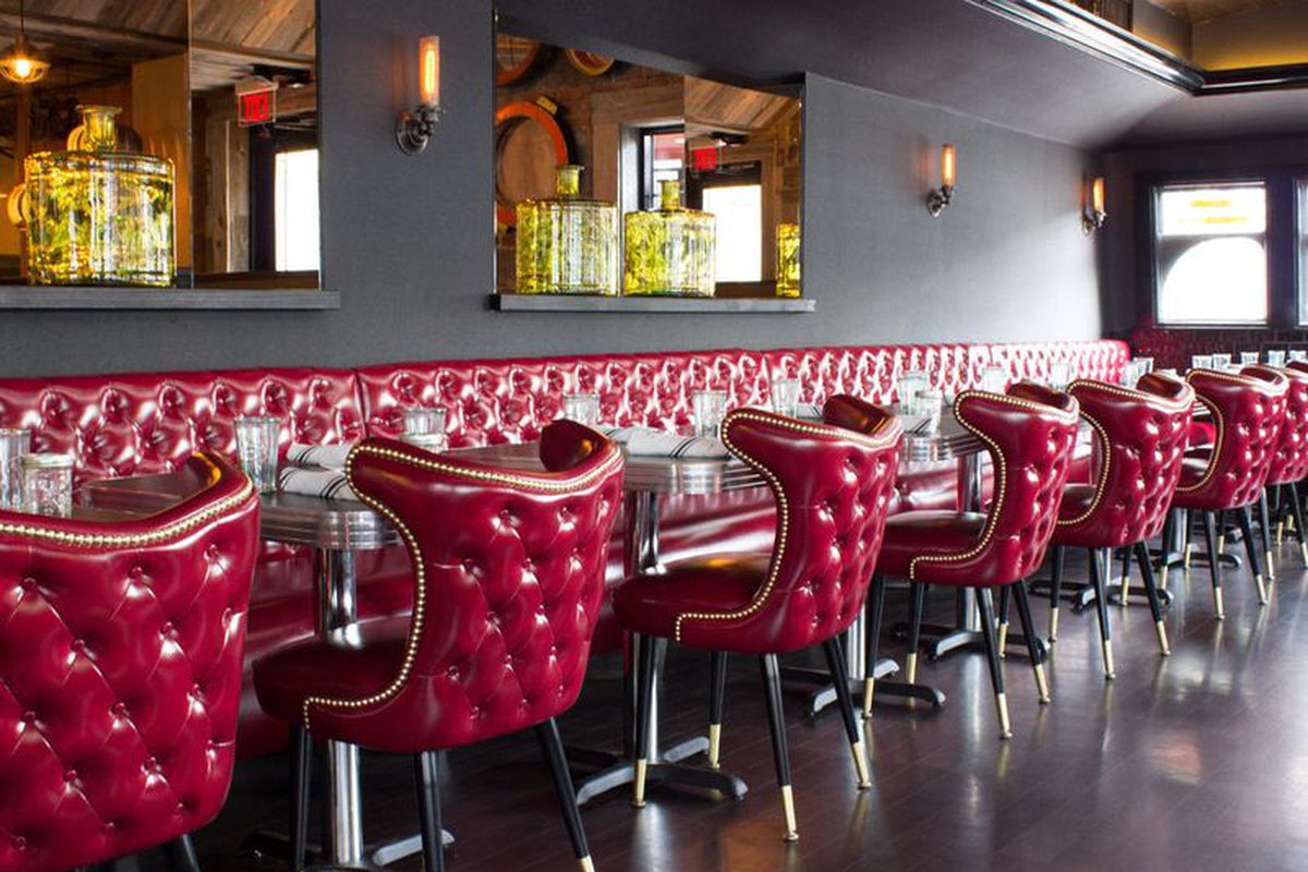 Interior of a restaurant, featuring shiny red leather chairs and banquettes, a dark gray wall, and mirrors.