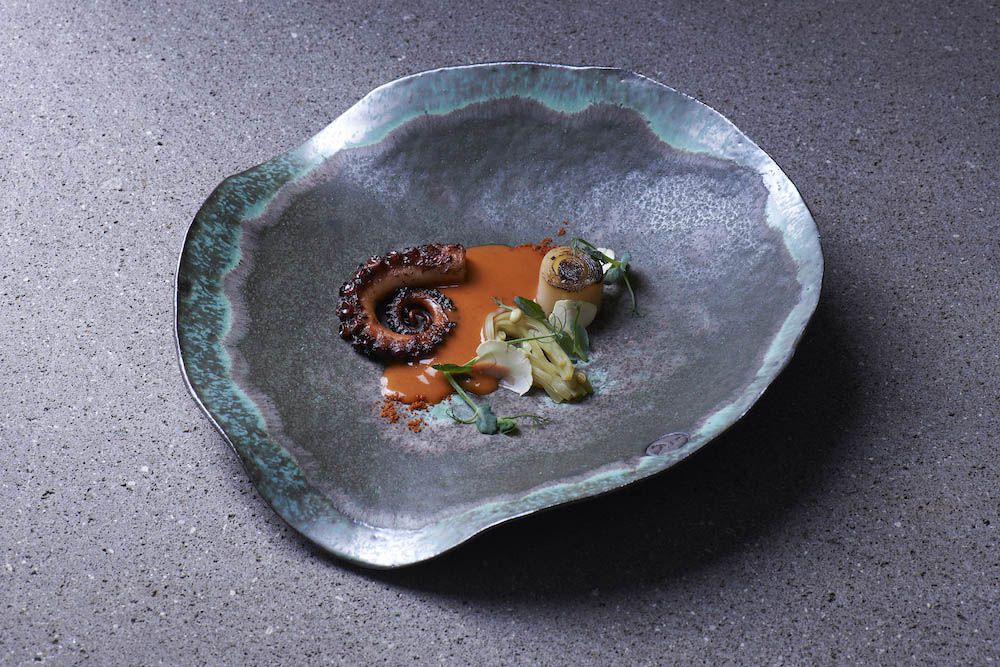 A large plate that looks like a sea shell, holding octopus and vegetables in a small pool of sauce.