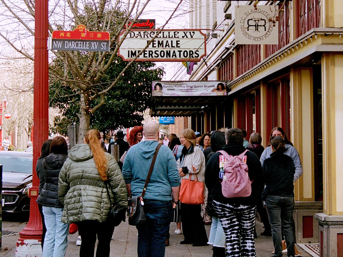 A line of people wait on the sidewalk outside Darcelle XV under a “Female Impersonators” sign.