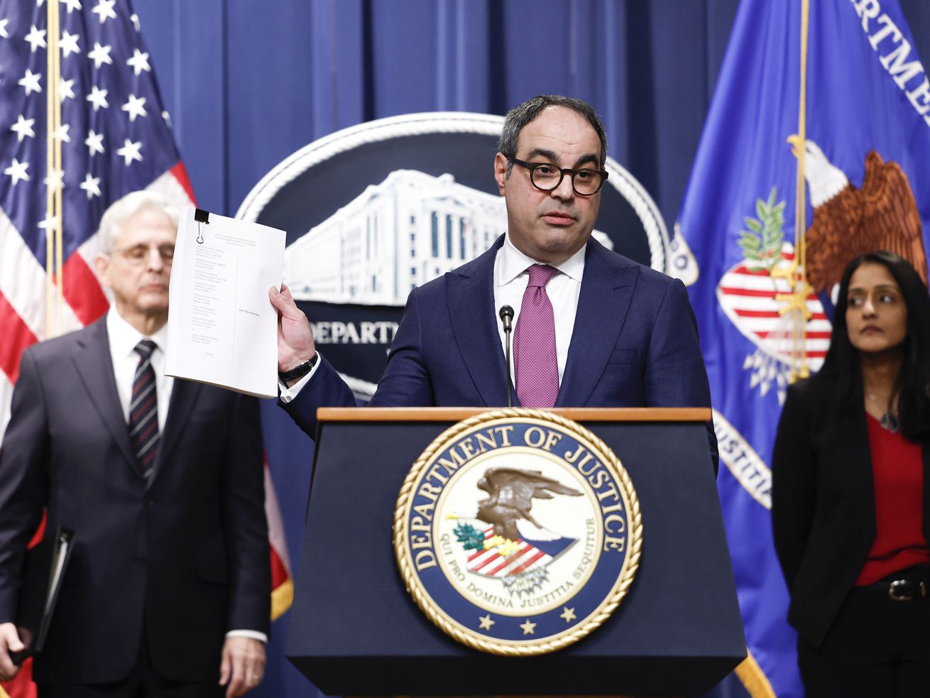 Jonathan Kanter holds up a document as he speaks alongside Attorney General Merrick Garland and Associate Attorney General Vanita Gupta at the Justice Department press podium with flags and a seal behind him. 