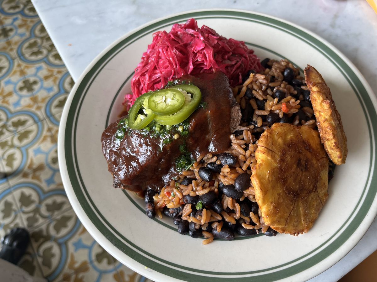 A plate of rice and beans topped with glazed barbecued ribs, fried tostones, and pink slaw. 
