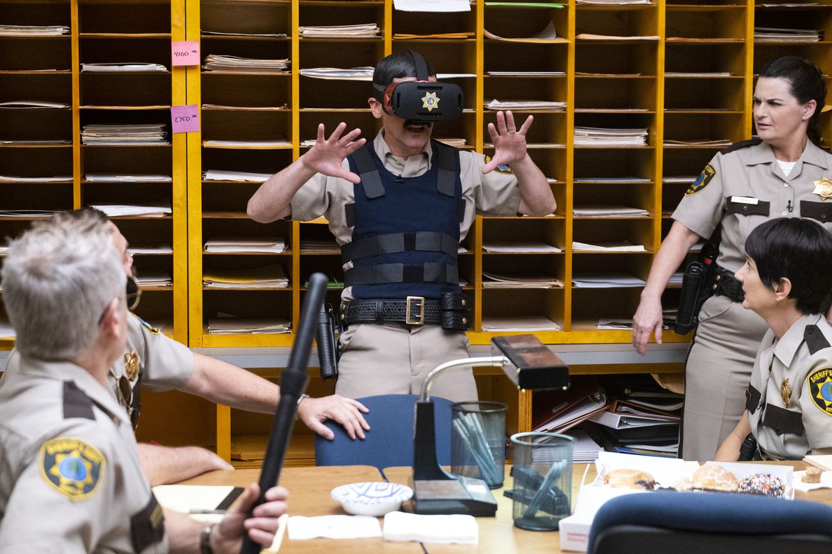 A Reno 911! character in a virtual-reality headset holds up his hands in excitement as other sheriff’s department employees watch.