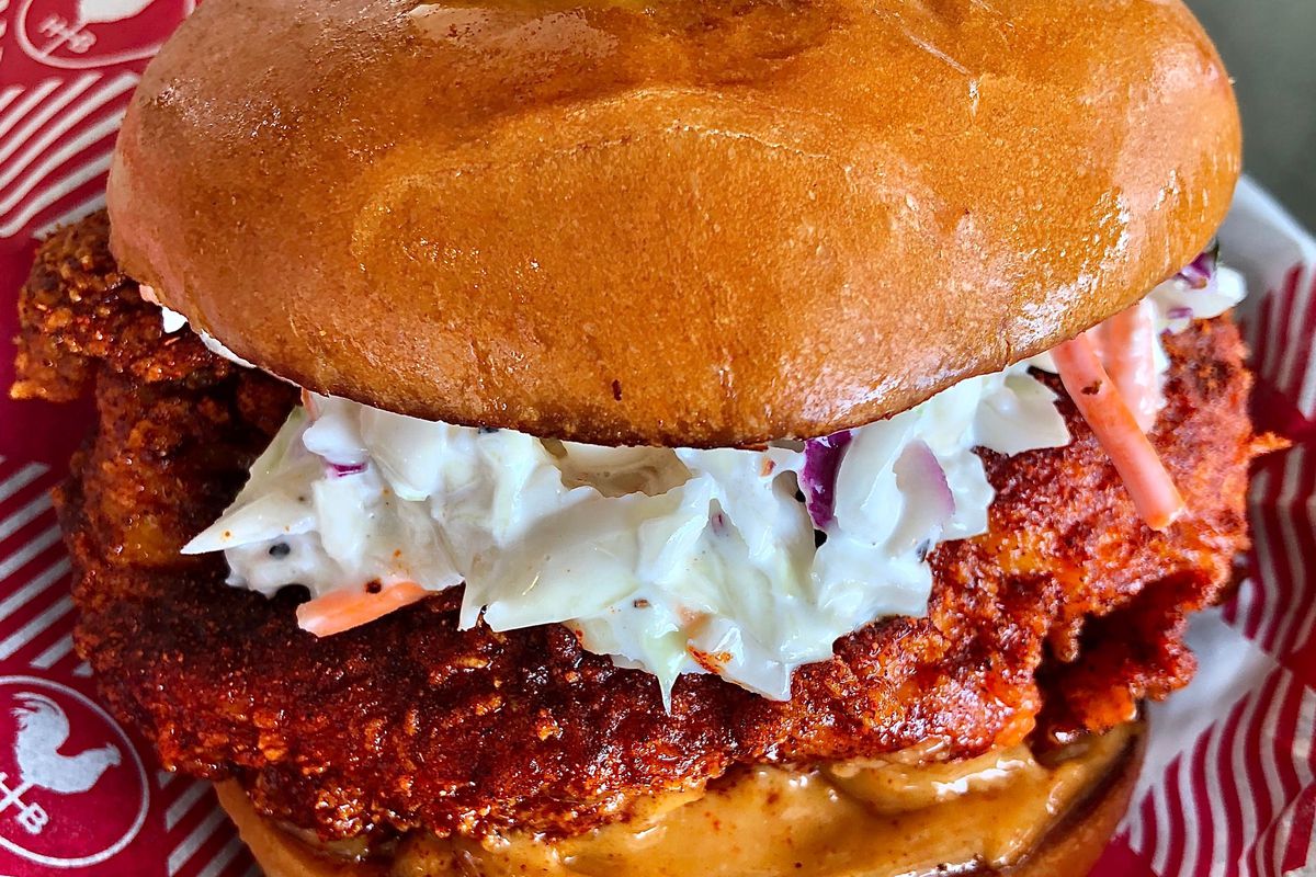 A hot chicken sandwich on red and white checked paper, topped with coleslaw and garnished with a pickle slice on a toothpick.
