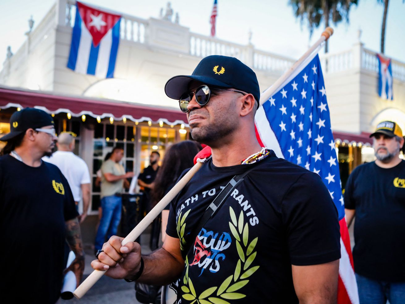 Tarrio wears a black shirt emblazoned with the Proud Boys logo, sunglasses, and a black baseball cap, and carries an American flag over his shoulder during a protest in Miami, Florida, on July 16, 2021.