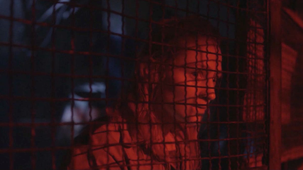 A woman stares through a chain-link fence bathed in red light in Virus-32.