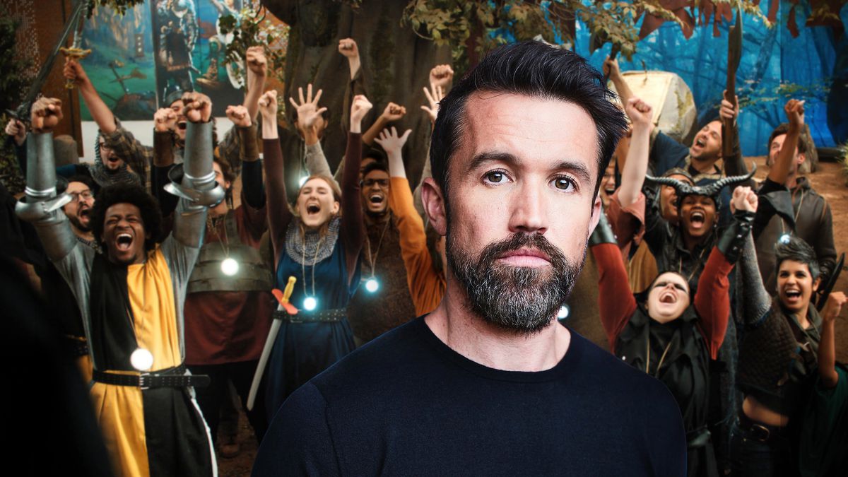 Actor Rob McElhenney looks to camera with a large cheering group of people in medieval costume cheering behind him