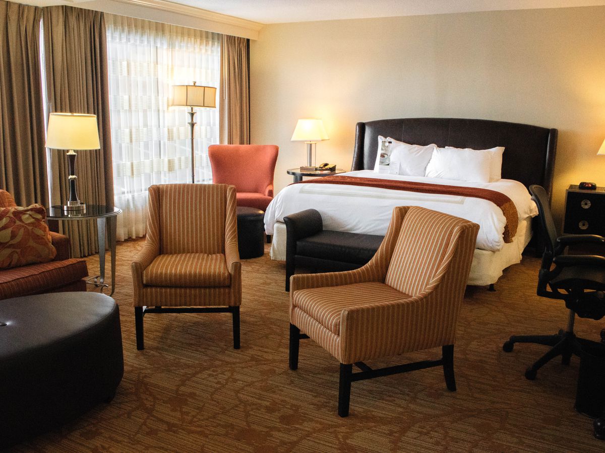 The interior of a hotel room. There is a bed, two arm chairs, a table, and multiple lamps.