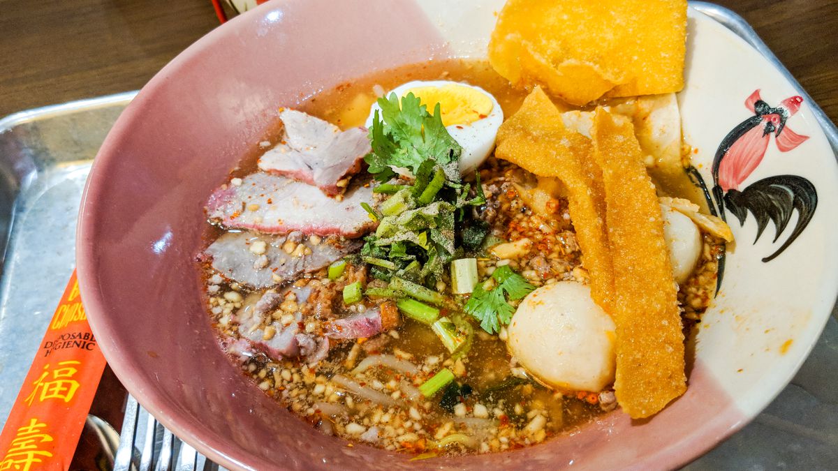 Soup in a Thai-style bowl decorated with a rooster. There are pink barbecue pork slices in the soup, as well as an egg, ground pork, ground peanuts, crispy wonton strips, and more. The bowl sits on a metal tray.