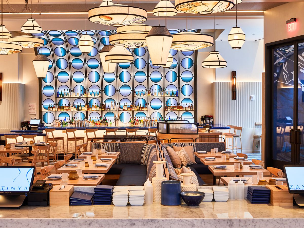 Linen-covered chandeliers hang from the ceiling over a dining room with blue banquettes and light wood tables, and a bar with spherical blue lights is visible in the background.