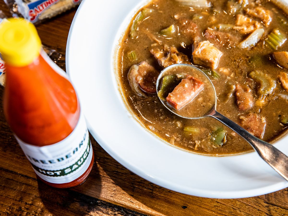 A bowl of sausage gumbo with a spoon in it and a bottle of hot sauce.
