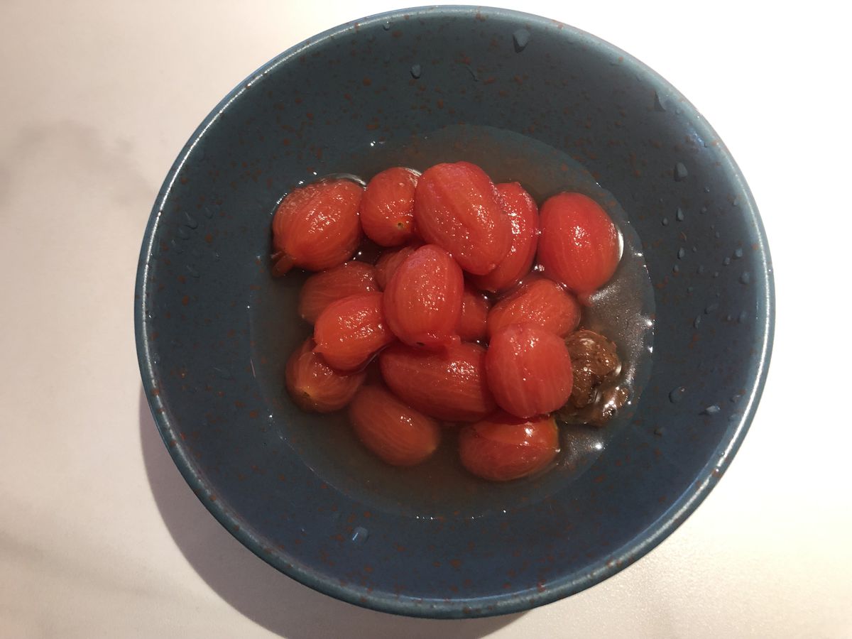 An overhead shot of a blue bowl with red tomatoes and brown plums in a clear liquid.