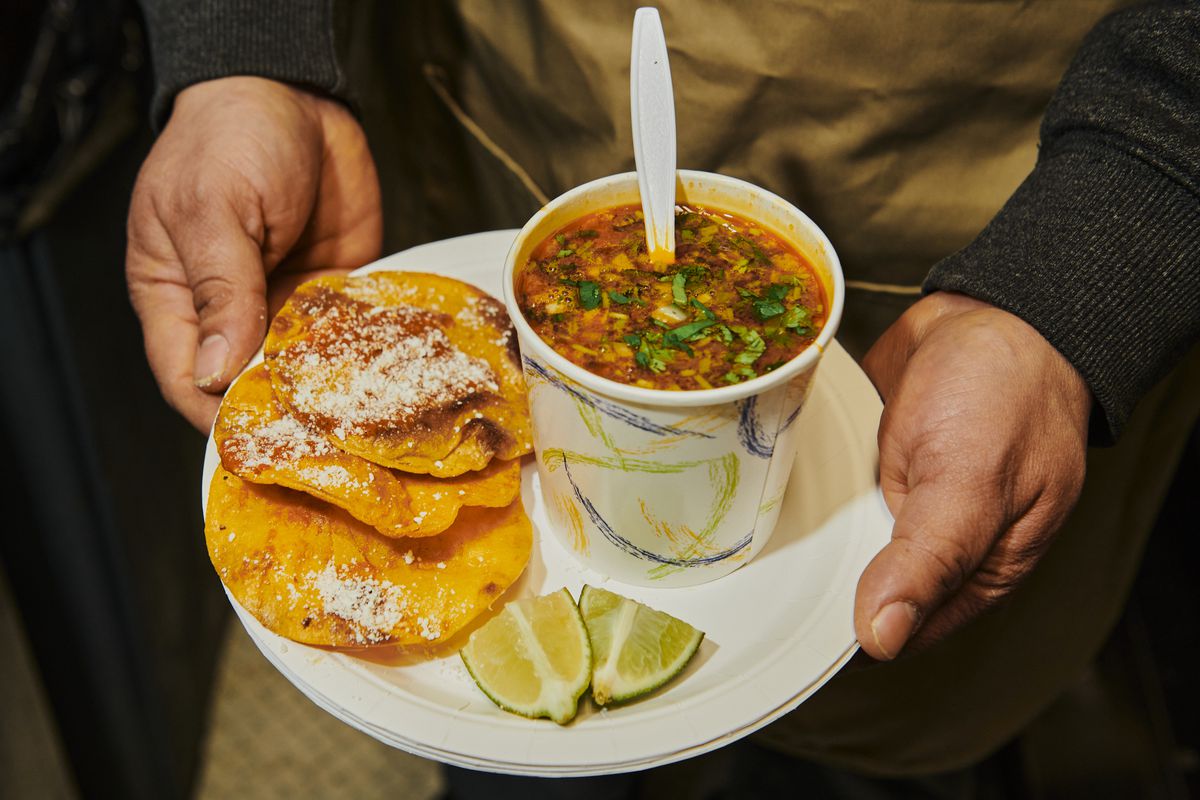 Two hands hold a disposable plate with three tostadas, a cup of brothy consomé, and two lime wedges.