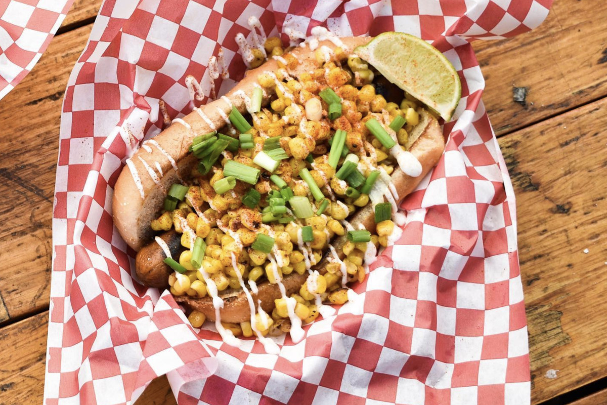 A vegan hot dog from Cycle Dogs loaded with toppings sits on top of checkered-patterned paper, with a side of lime.