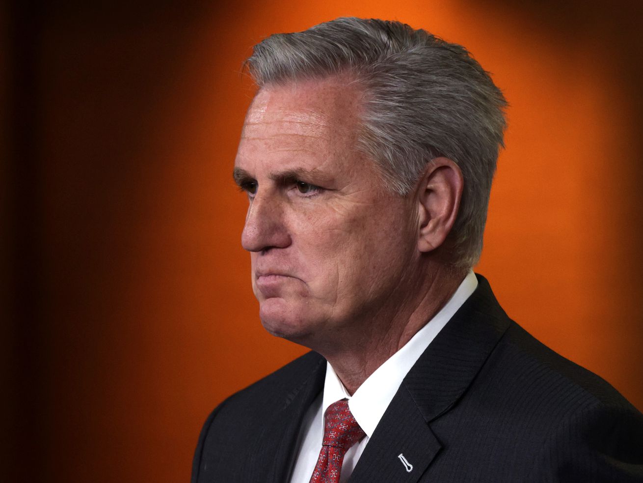 Kevin McCarthy, in a black suit and red tie, looks to the left with a scowl on his face, against a dark orange background.