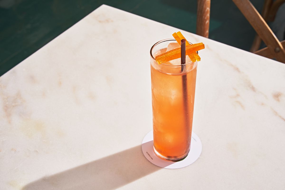 An orange cocktail is filled in a slender glass with a black straw, sitting on a marble counter top.