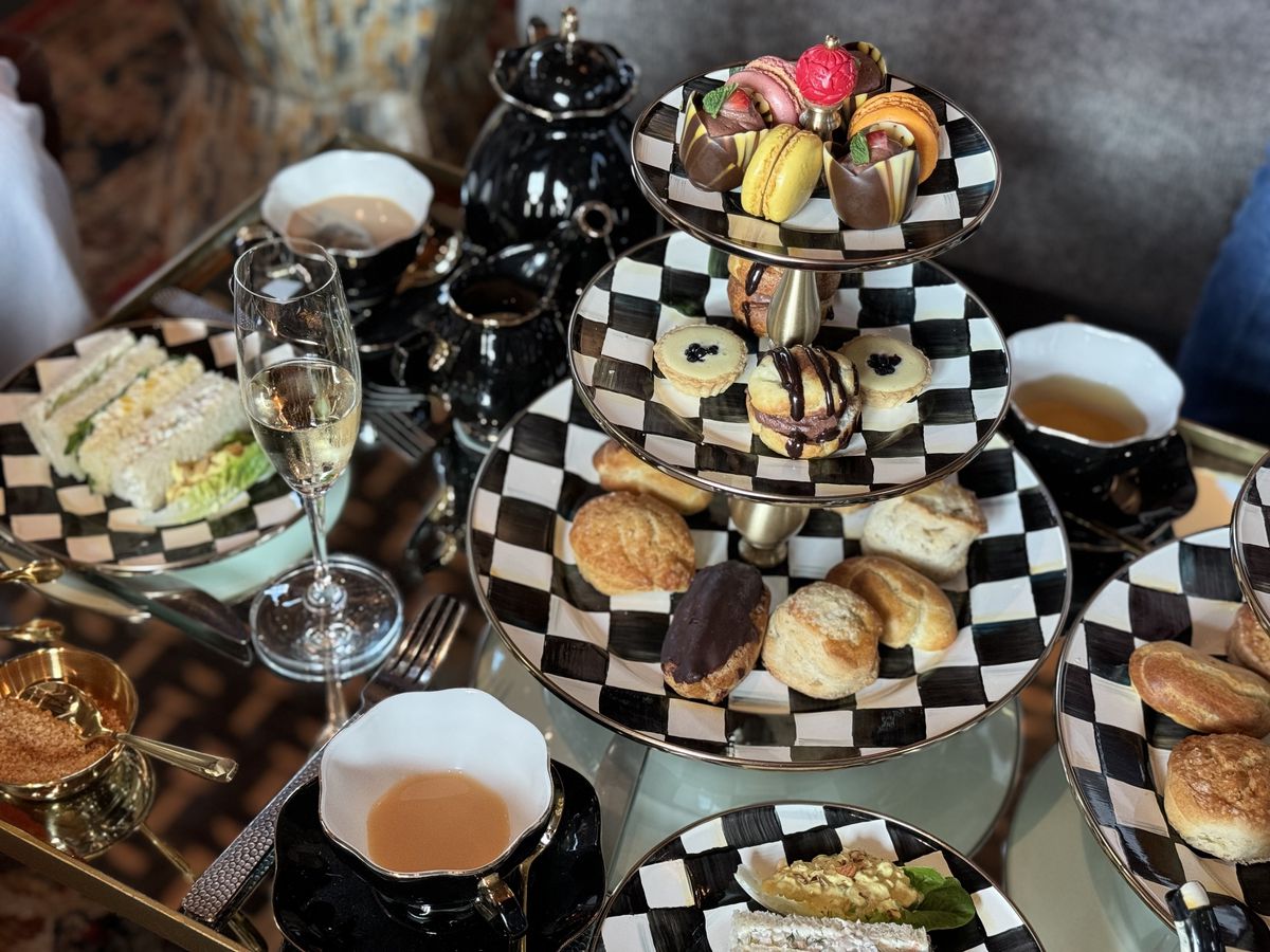 A checkered tea set and tower plate holds scones, pastries, mugs of tea, and sandwiches.