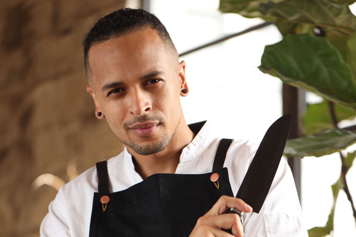 A man with facial hair, a white chef’s jacket, black apron, and a black knife posing intensely at the camera.