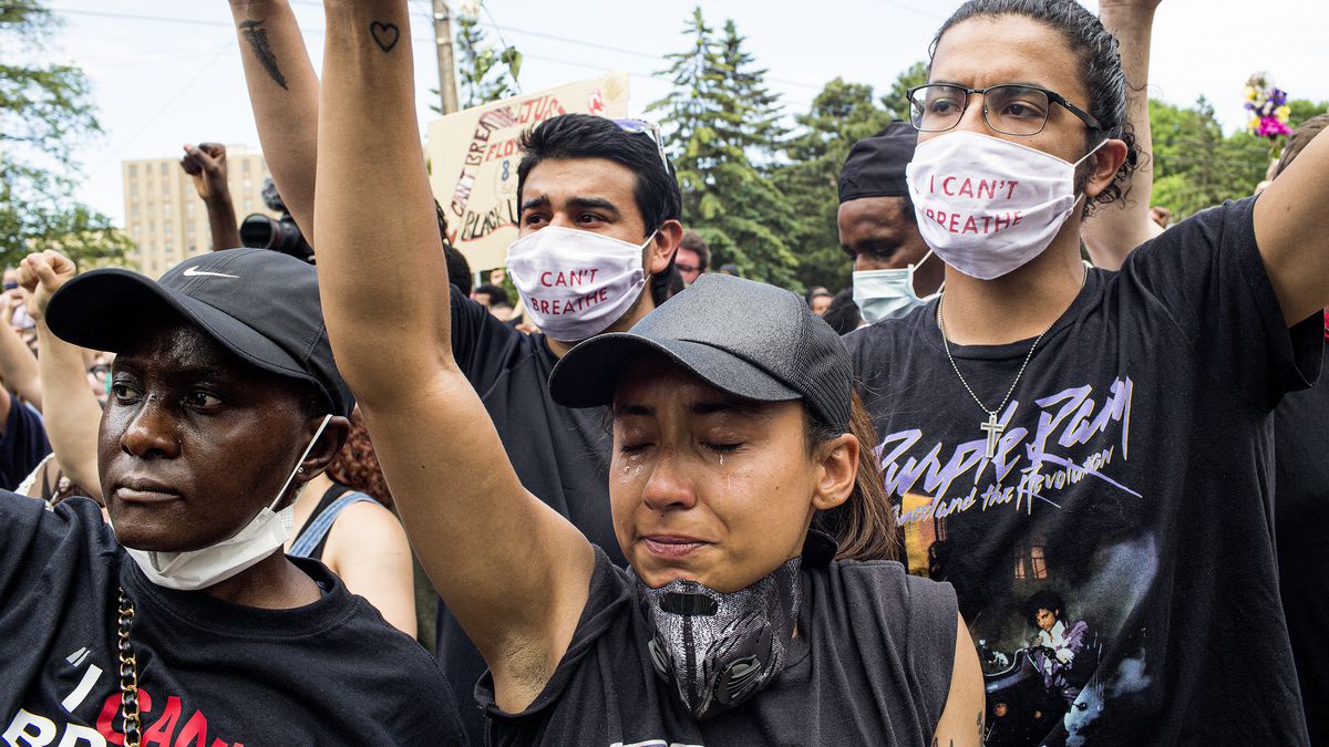 A group of people, men and women, of all ethnicities, dressed in black and wearing masks, raise their fists.