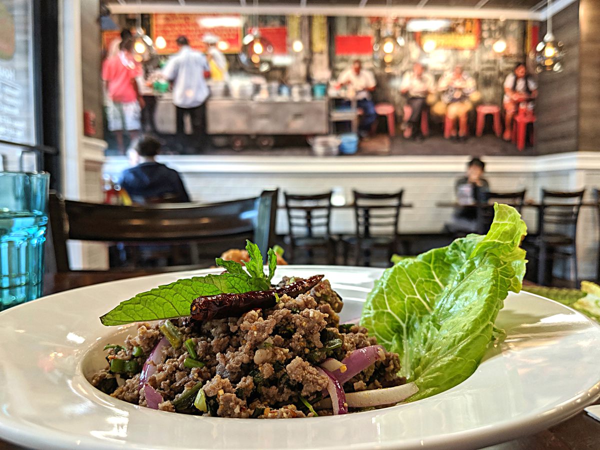 A pile of seasoned ground beef studded with sliced red peppers and toasted rice sits on a restaurant table with a colorful photo of Thailand in the background.