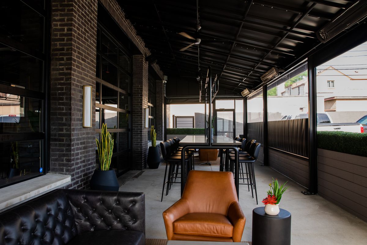 A long table and chairs in the background, a brown leather seat and a black leather sofa and green plant accents under an enclosed patio