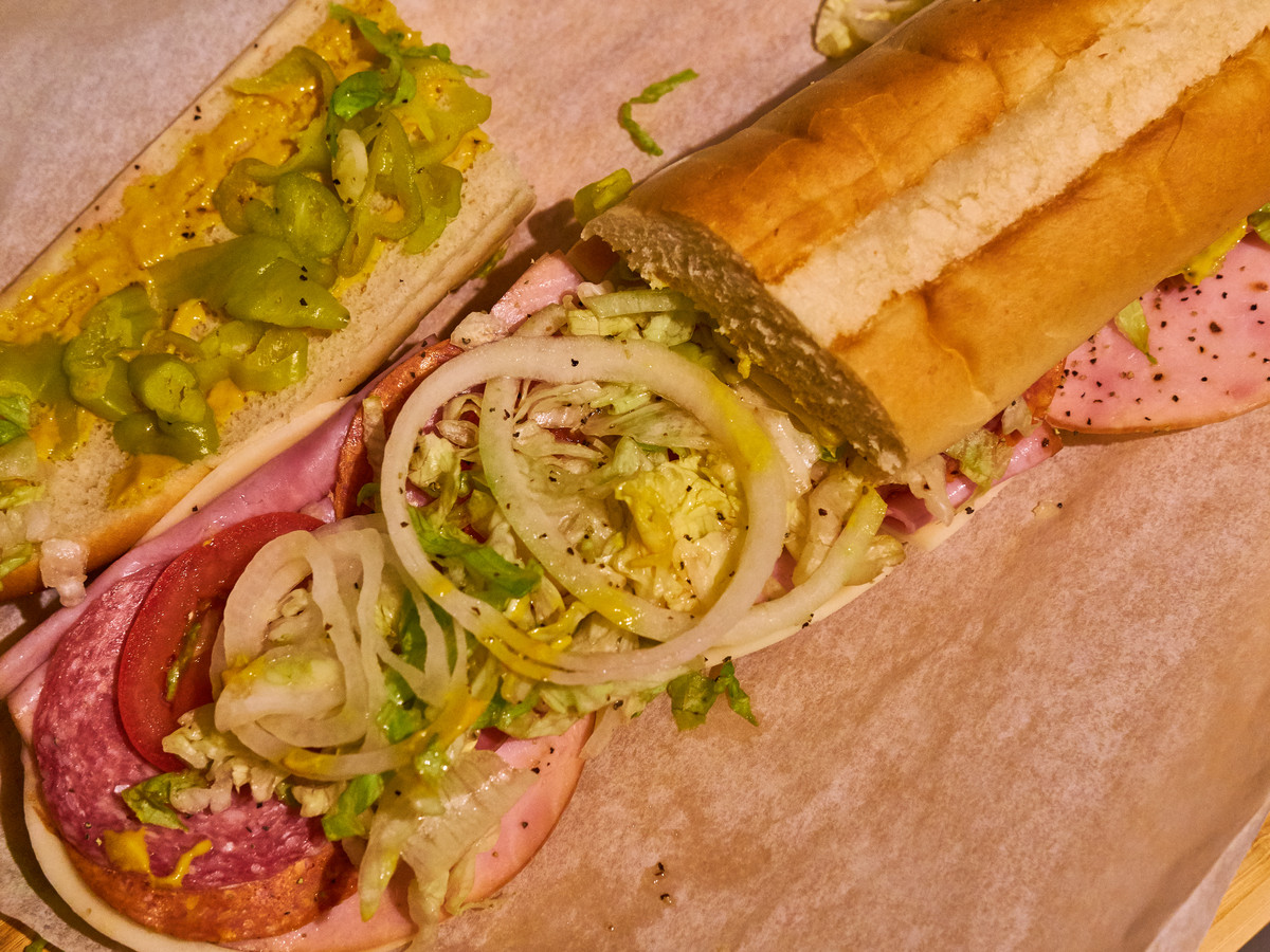 A footlong sub sandwich with deli meats, onions, greens, and peppers