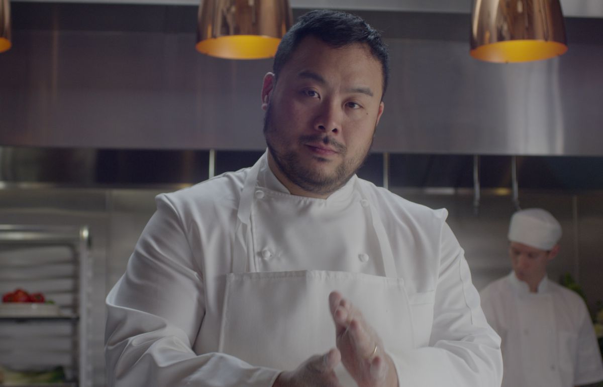 Chef David Chang wearing all white chef’s coat and apron, standing in a restaurant kitchen.