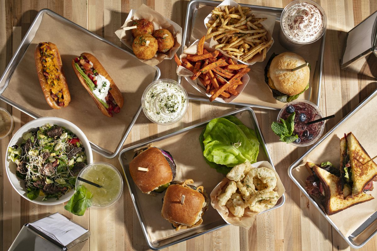An overhead photograph of metal trays lined with food, including hot dogs, salads, burgers, onion rings, and fries