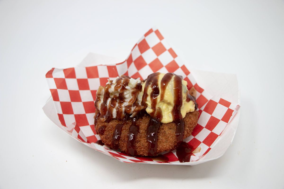 Two balls of fried food are placed in a red checkered serving container.  On top are servings of coleslaw, potato salad, and barbecue sauce. 