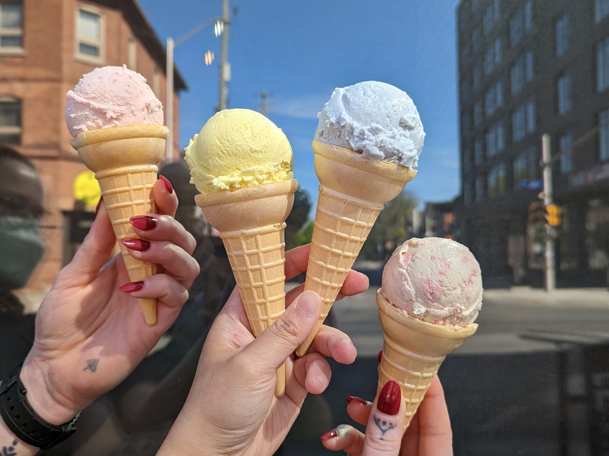 Hands hold four brightly colored ice cream cones in front of a street on a clear day. 