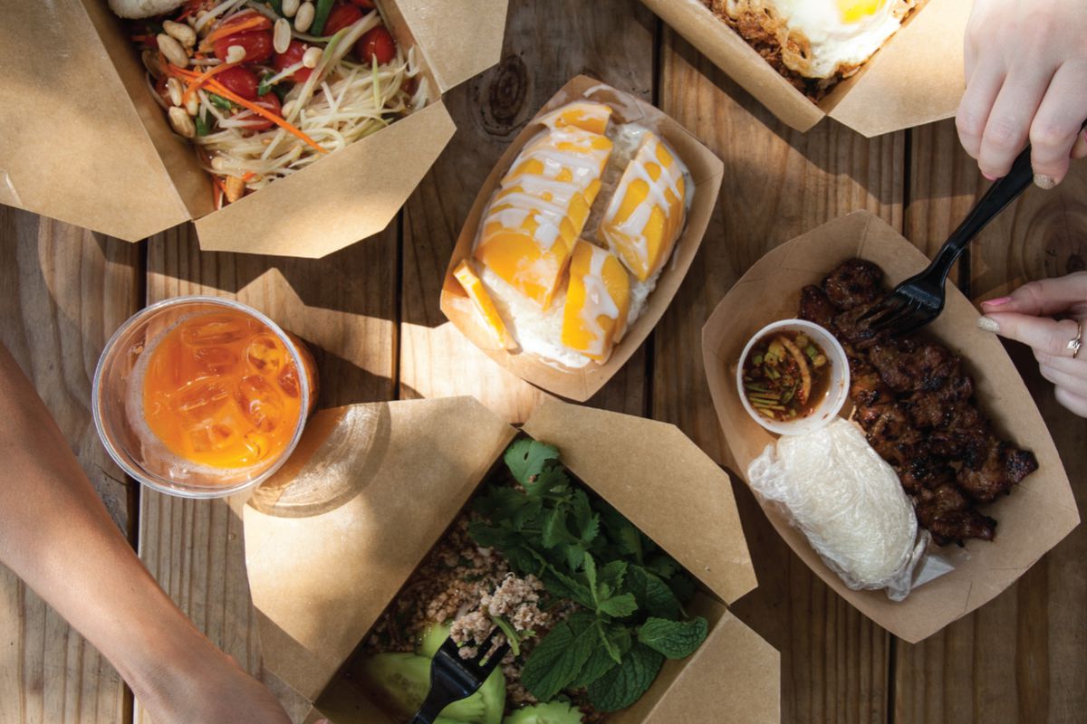 An array of Thai dishes in cardboard containers on a table with people reaching into them.