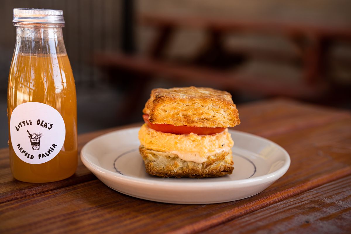 A place with a biscuit sandwich of thick egg and a slice of tomato next to a clear bottle of orange liquid with a label reading Little Ola’s Arnold Palmer.