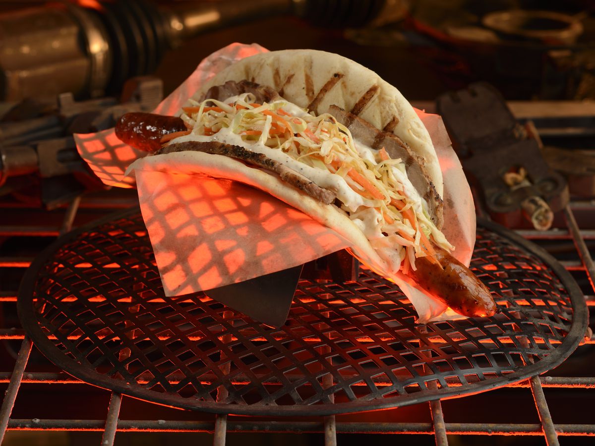 A ronto wrap (sausage pita sandwich)in a cardboard boat on a grill rack as if lit by the light of a fire.