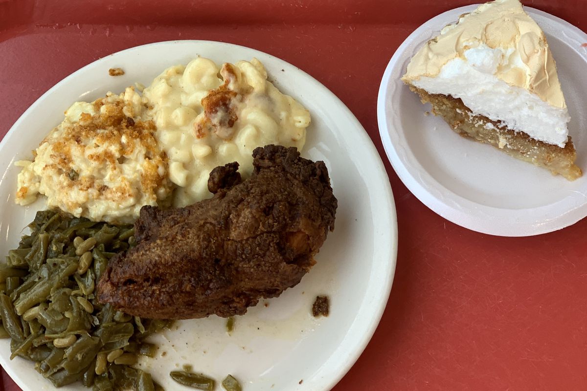 A red cafeteria tray on a wood table. Left plate topped with dark fried chicken, greens, cauliflower casserole. Right plate has slice of chess pie.