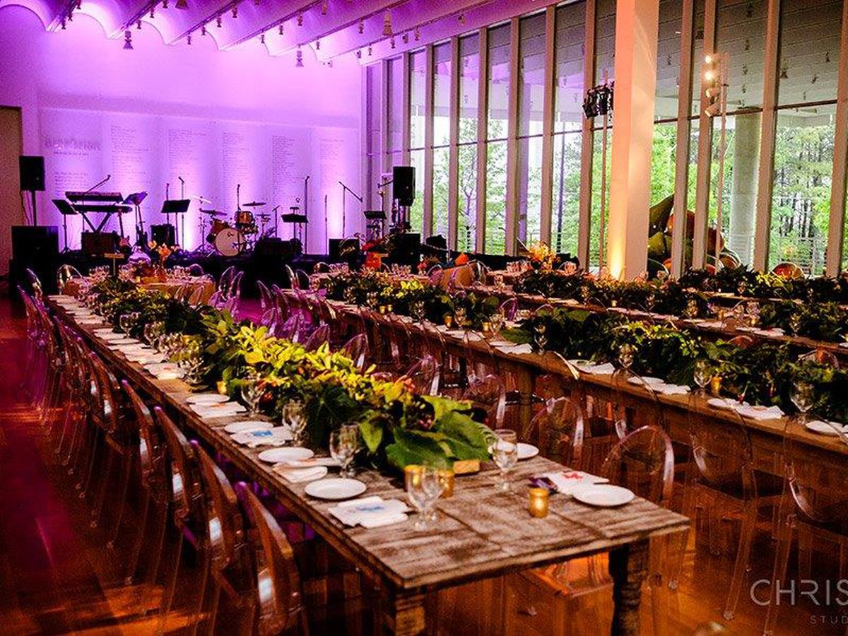 A large wedding reception event space with long tables and a multitude of chairs. The tables have elaborate floral centerpieces and dishes on them. There is a stage at the end of the room which is lit in purple stage lighting. 