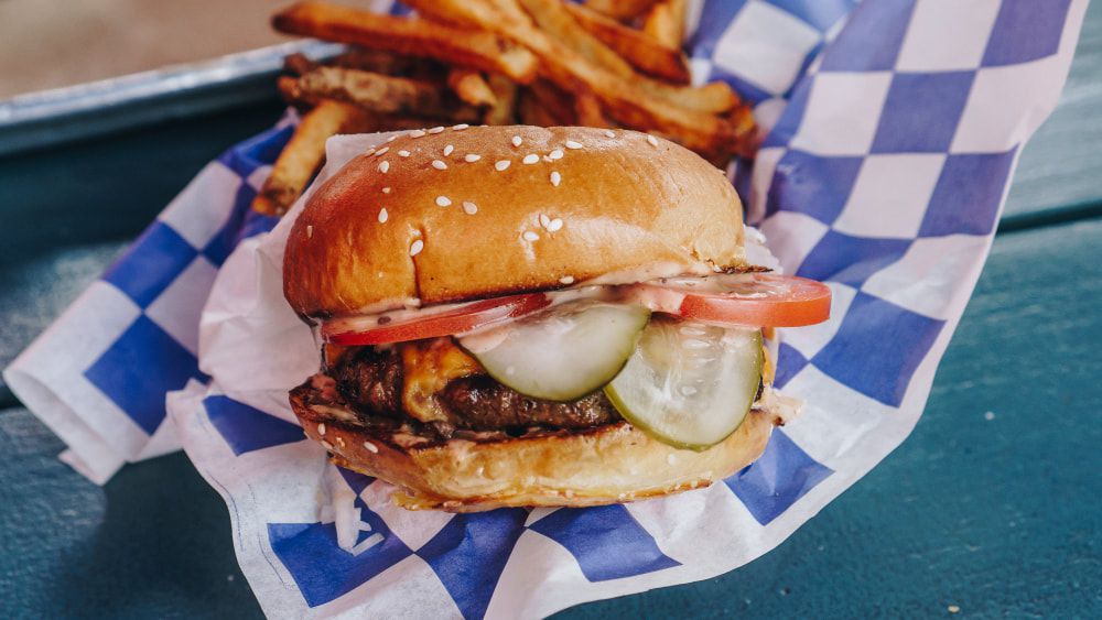 A burger with pickles and tomatoes on a seasame bun sits on a blue and white checkered paper.