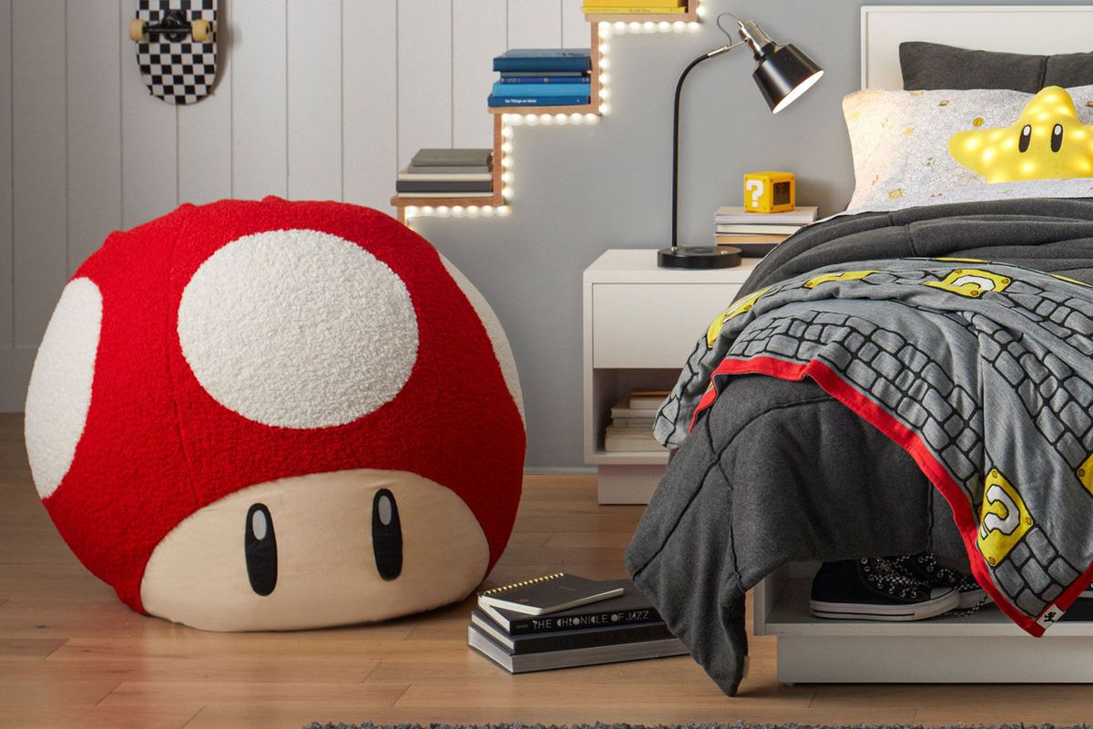 A photo of a bedroom that’s adorned with Super Mario-related home decor from Pottery Barn. This includes a large bean bag chair based in design on the Mushroom collectible item. There’s also a Mario-themed throw, multiple pillows, and a wall-mounted LED in the shape of a Mushroom power up.