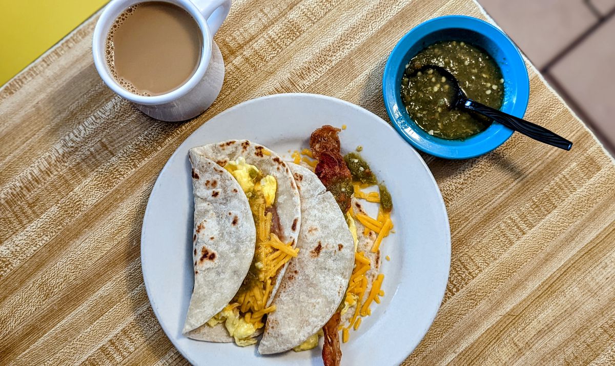A white plate with two tacos with eggs and shredded orange cheese and one with a bacon strip next to a mug of coffee and a blue saucer of condiments on a table.