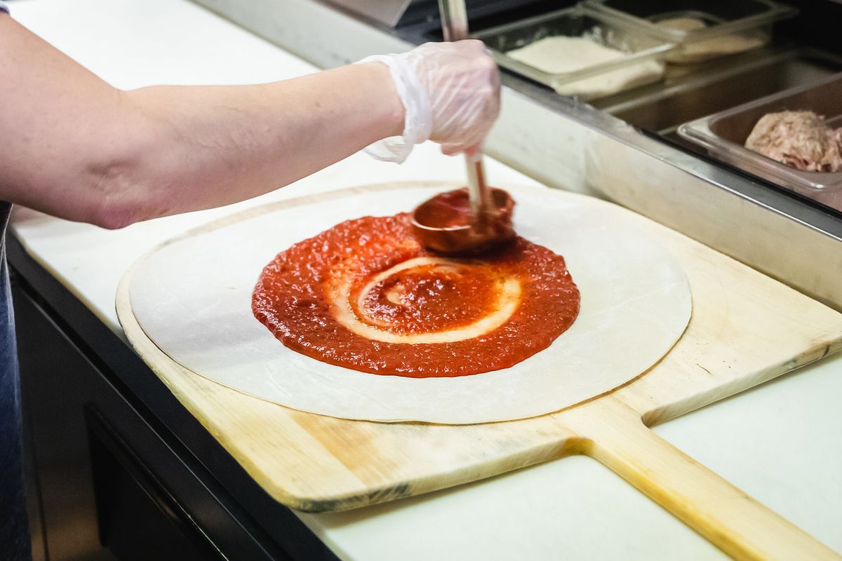 An arm holding a metal ladle to apply red sauce to a pizza crust.