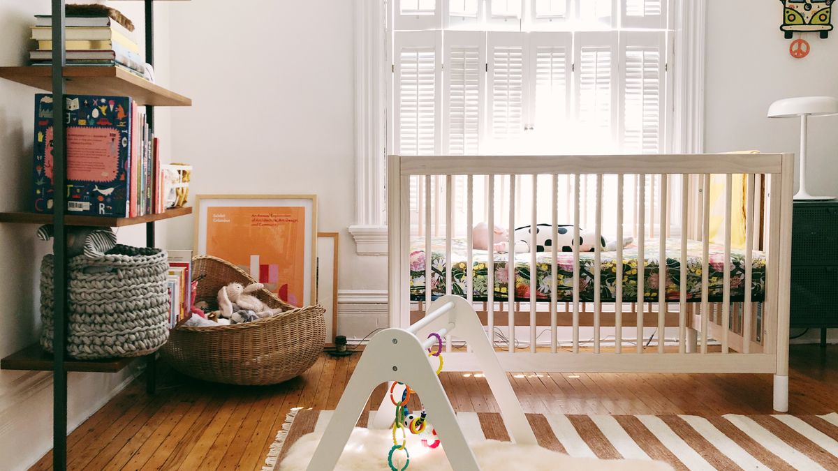 A white-walled room contains a white crib with a baby in it, a bookshelf holding books and a basket, a brown and white striped area carpet on a hardwood floor, and a white baby’s jungle gym.