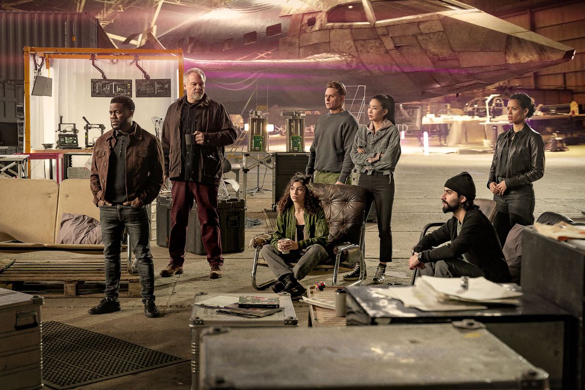 The cast of Lift, including Kevin Hart and Gugu Mbatha-Raw, stand around in a hangar with a fake CGI plane behind them