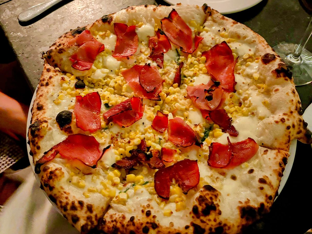 A pizza with a blistered crust and topped with soppressata and corn on a table.
