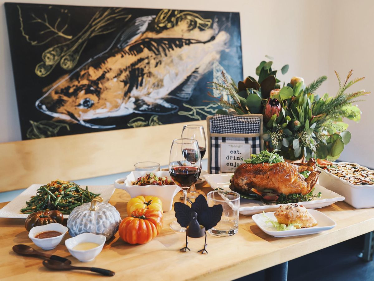 A table covered with a roasted duck, gourds, sauces in tureens, a turkey figurine, and two glasses of red wine, with a painting of a catfish in the background