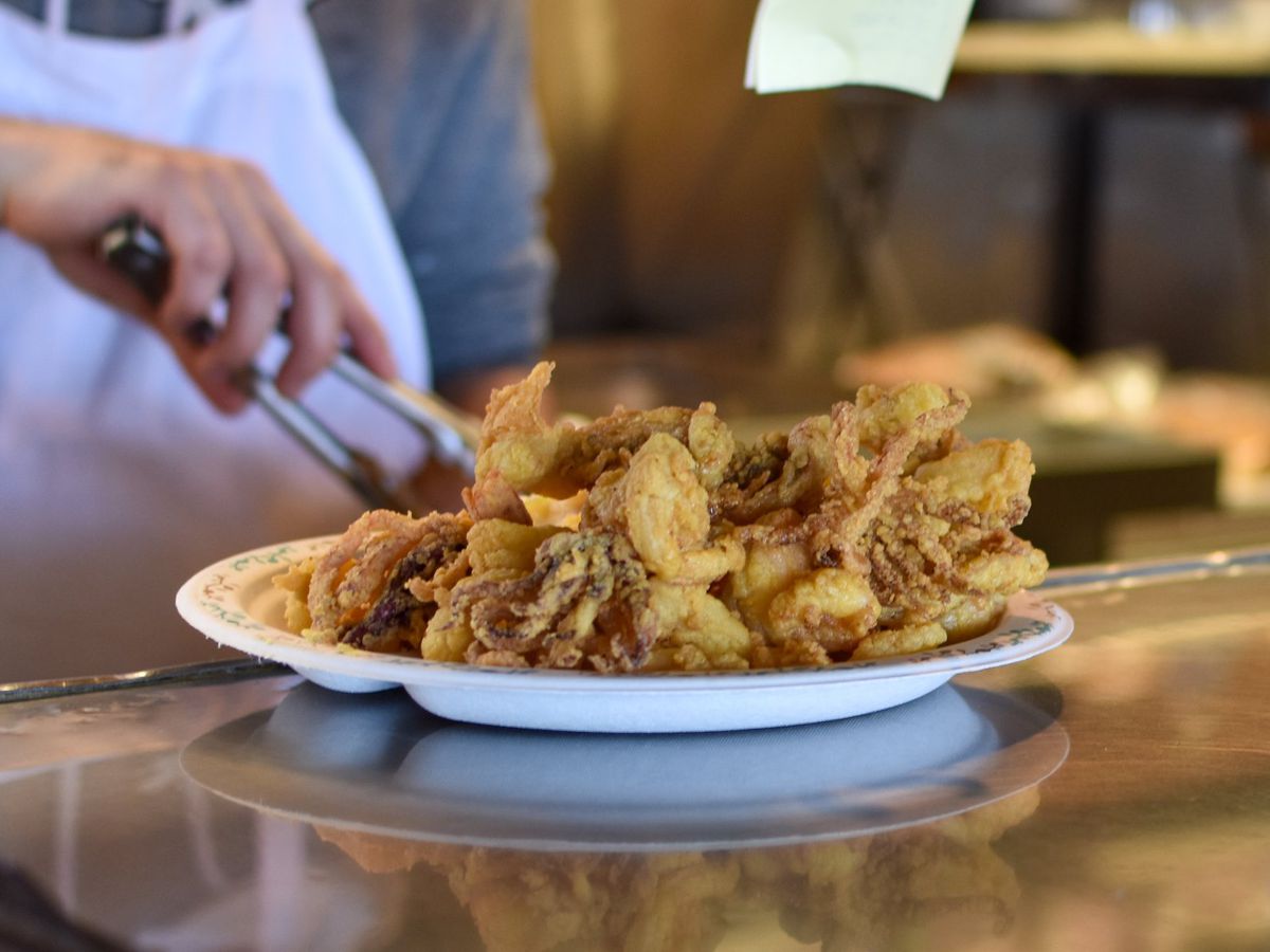 A hand uses metal tongs to grab some fried seafood from a white paper plate, which is loaded high with the seafood and sits on a counter.