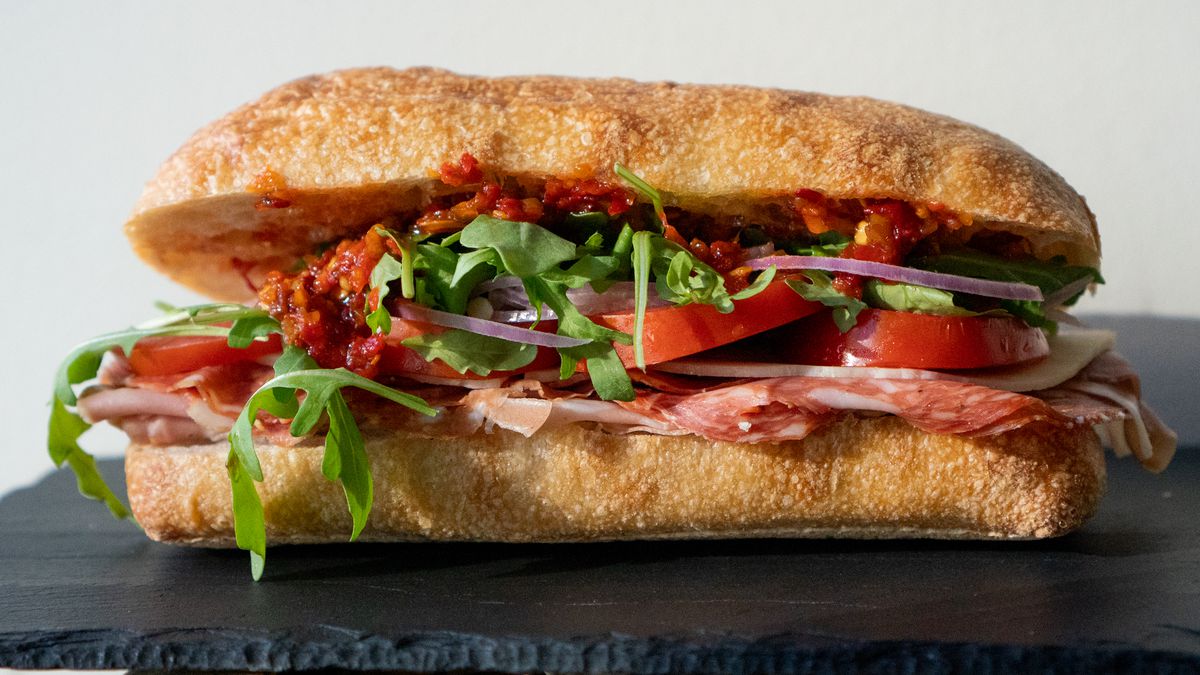 Slices of deli meats, arugula, tomatoes, and a bright red pepper spread are sandwiched between two long pieces of ciabatta.