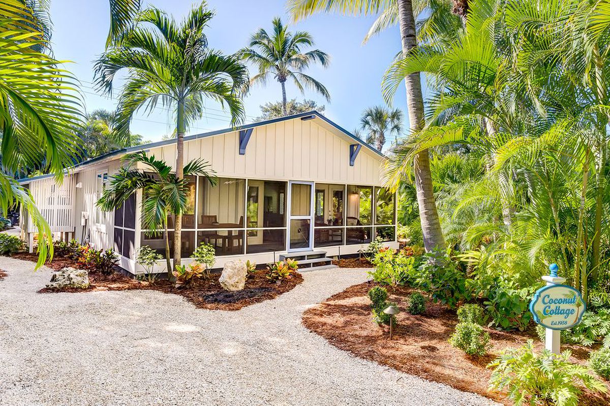 Exterior shot of tan-colored beach cottage with gable roof trimmed in blue with palm trees surrounding it. 