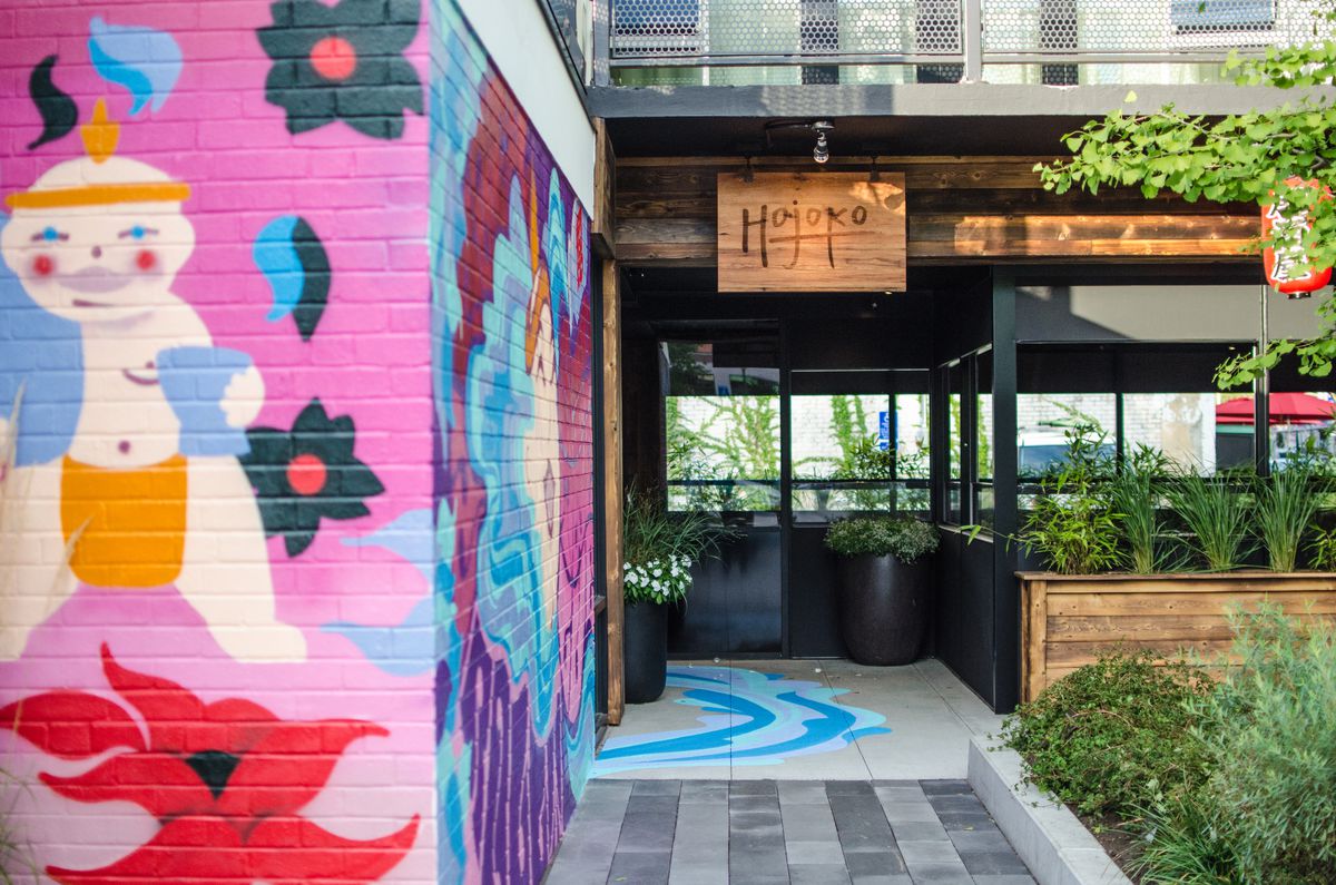 A colorful Japanese-inspired mural leads to the door of a restaurant with wooden signage reading “Hojoko.” There’s some greenery around the exterior.