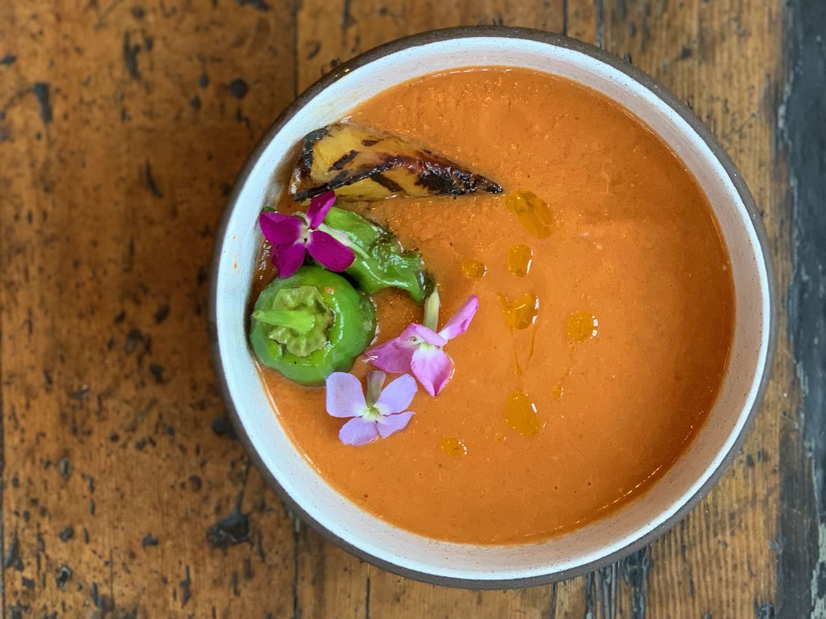 An bowl of orange-red gazpacho topped with grilled peach and pappers.